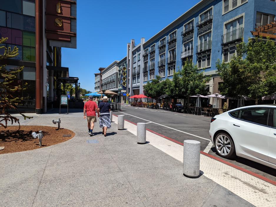 Photo of small street with people walking on sidewalk surrounded by large buildings and trees