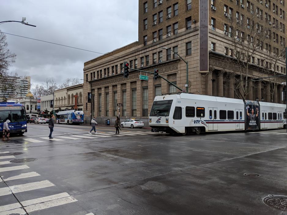 S2-C6.2-G3_DowntownSJ.jpg A photo of a light rail train traveling downtown near a VTA bus and people crossing the street.