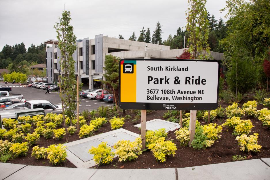Photo of sign for park and ride area in front of the parking lot and garage with cars parked