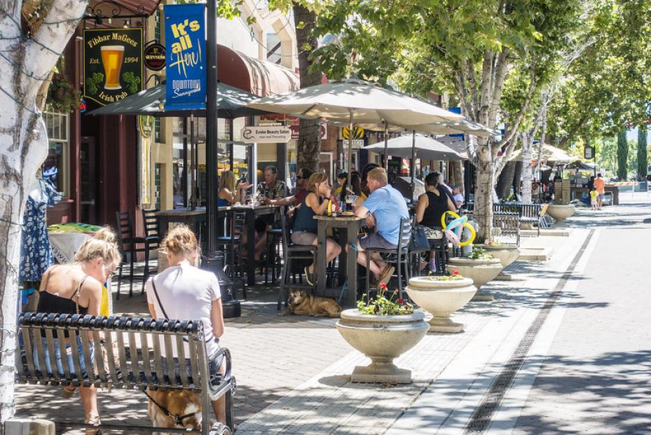 Photo of people sitting outside restaurants on benches and at tables under trees and umbrellas